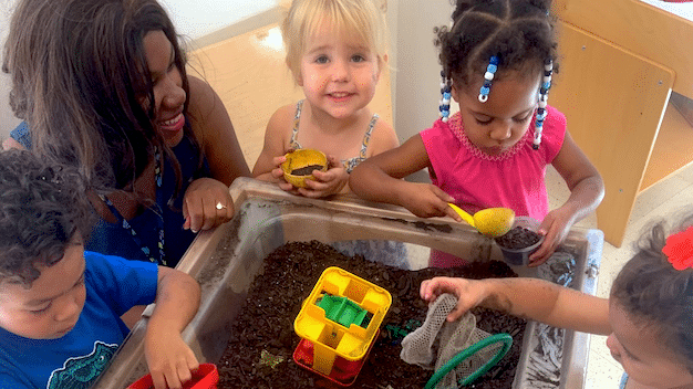 Children play with toys in dirt with teacher in classroom