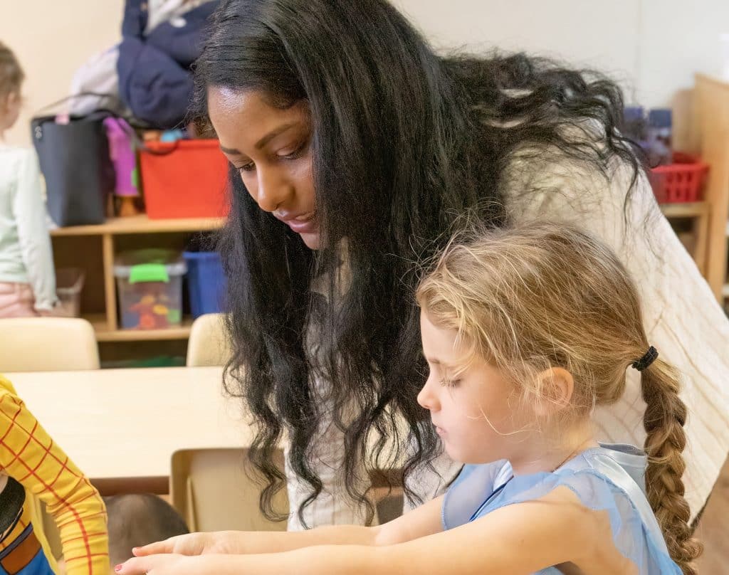 Child and educator peer over a bin of materials as child adds some to the bin.