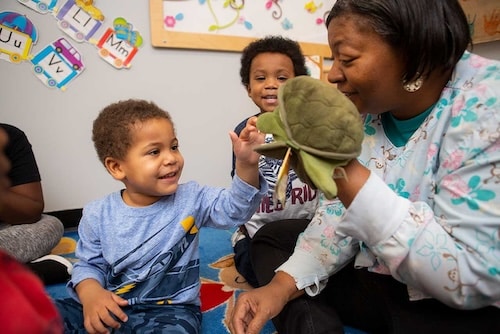 Educator sits with two children and with a turtle puppet on her hand. One child reaches for the puppet while smiling.