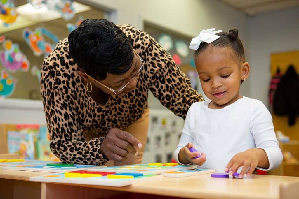 Child and educator in classroom. Child places shape blocks on a table as educator looks on.